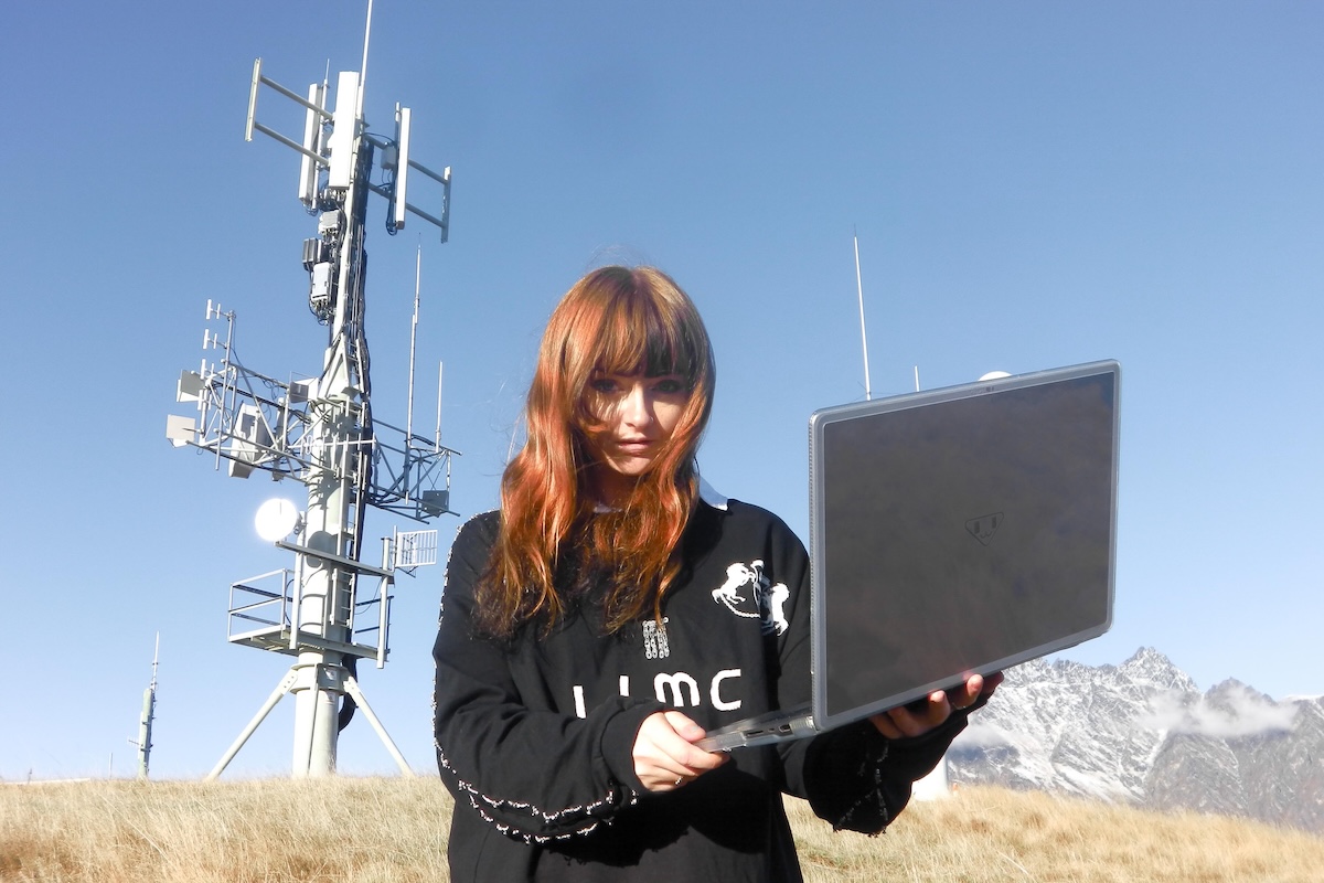 Australian producer Ninajirachi holding an open laptop outdoors with a communications tower and snowy mountain peaks in the background.